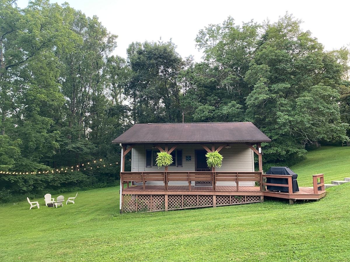 A cozy guest house is set back from the property line, surrounded by lush greenery. A spacious deck features two hanging planters and an outdoor grill. Nearby, an arrangement of four white chairs creates a relaxed seating area on the grass.