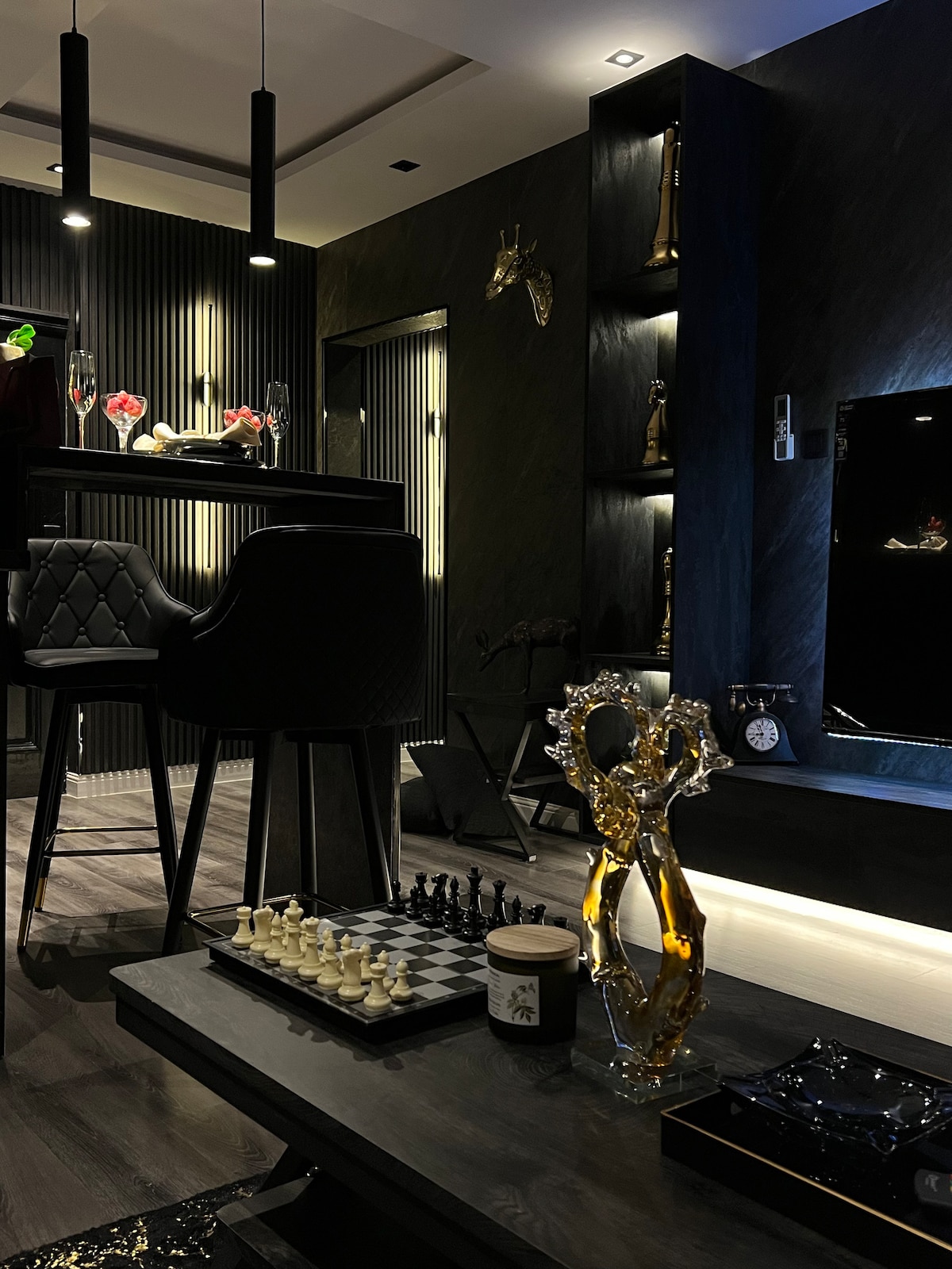 The living area features a modern black and gray color palette, with a stylish bar counter and high chairs. A chess set is displayed on a sleek table, along with decorative items on the shelves. Soft lighting creates an elegant ambiance throughout the space.