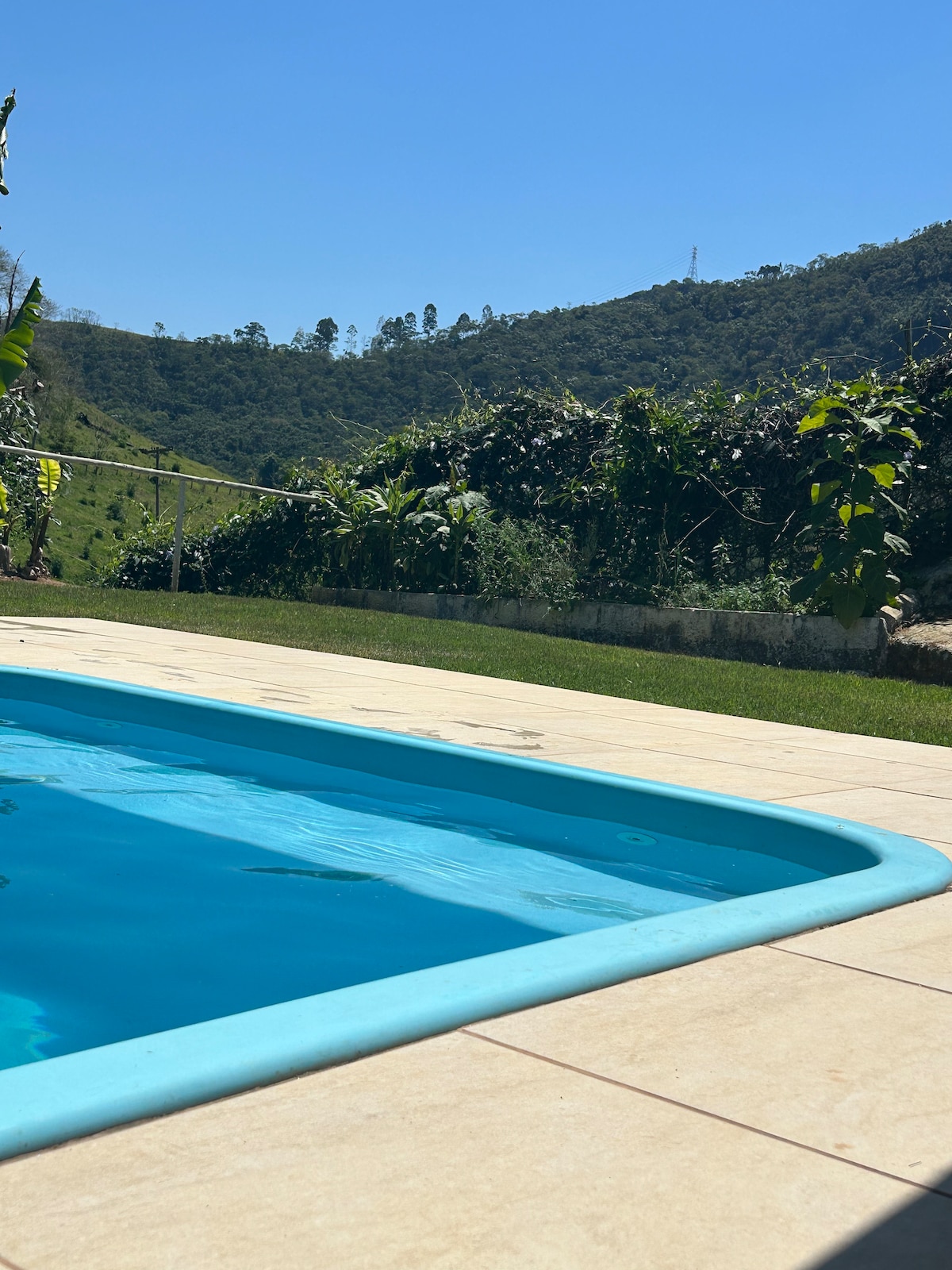 A refreshing pool is situated amidst lush greenery, with hills rising in the background. The clear blue water contrasts with the light-toned tiles surrounding the pool. Sunlight bathes the area, highlighting the natural beauty of the landscape and the vibrant foliage.
