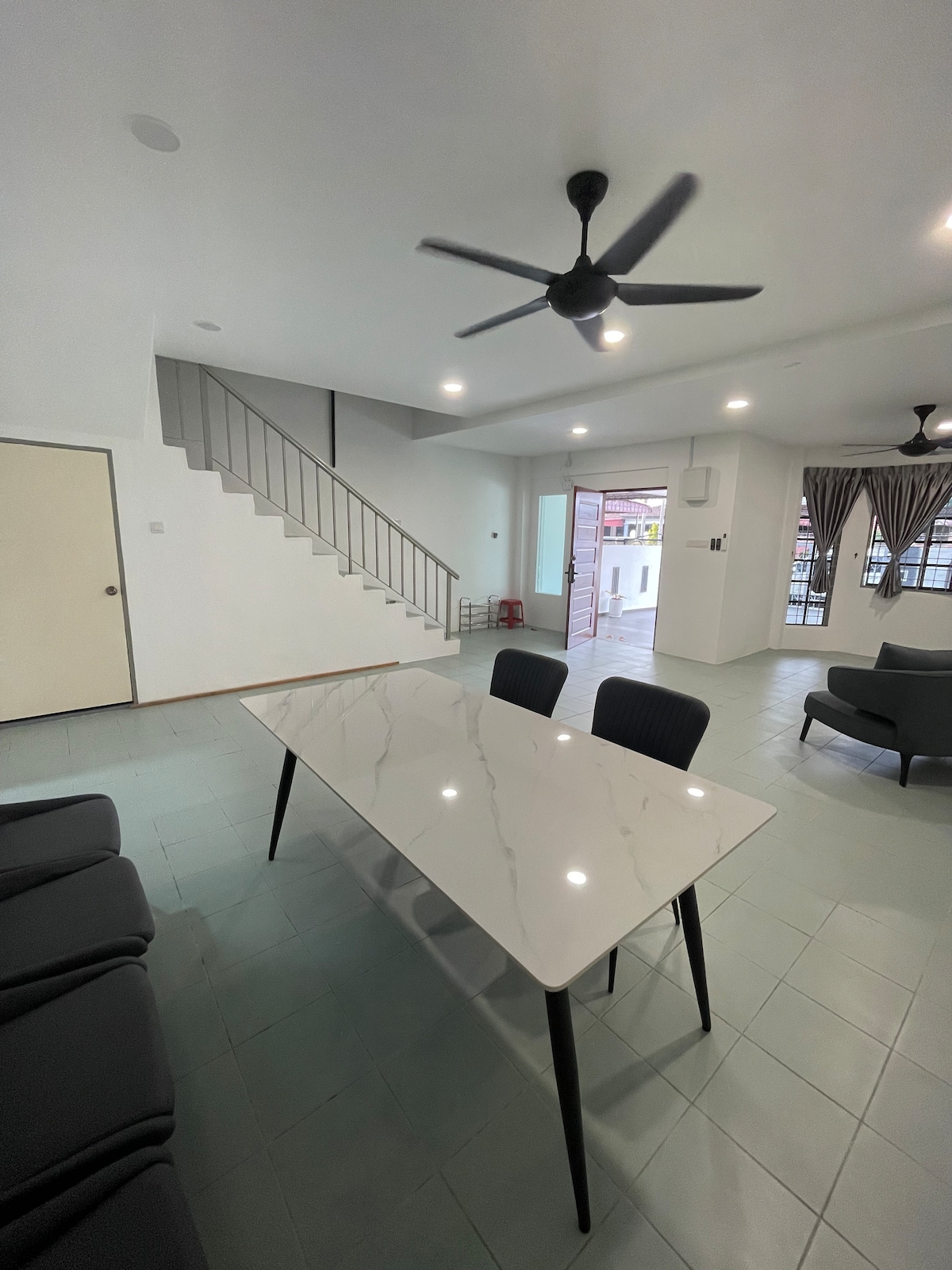 A modern dining area features a sleek marble table surrounded by four dark chairs. The open space includes a comfortable seating arrangement and is illuminated by natural light. A ceiling fan and light fixtures enhance the room's functionality. A staircase is visible in the background.