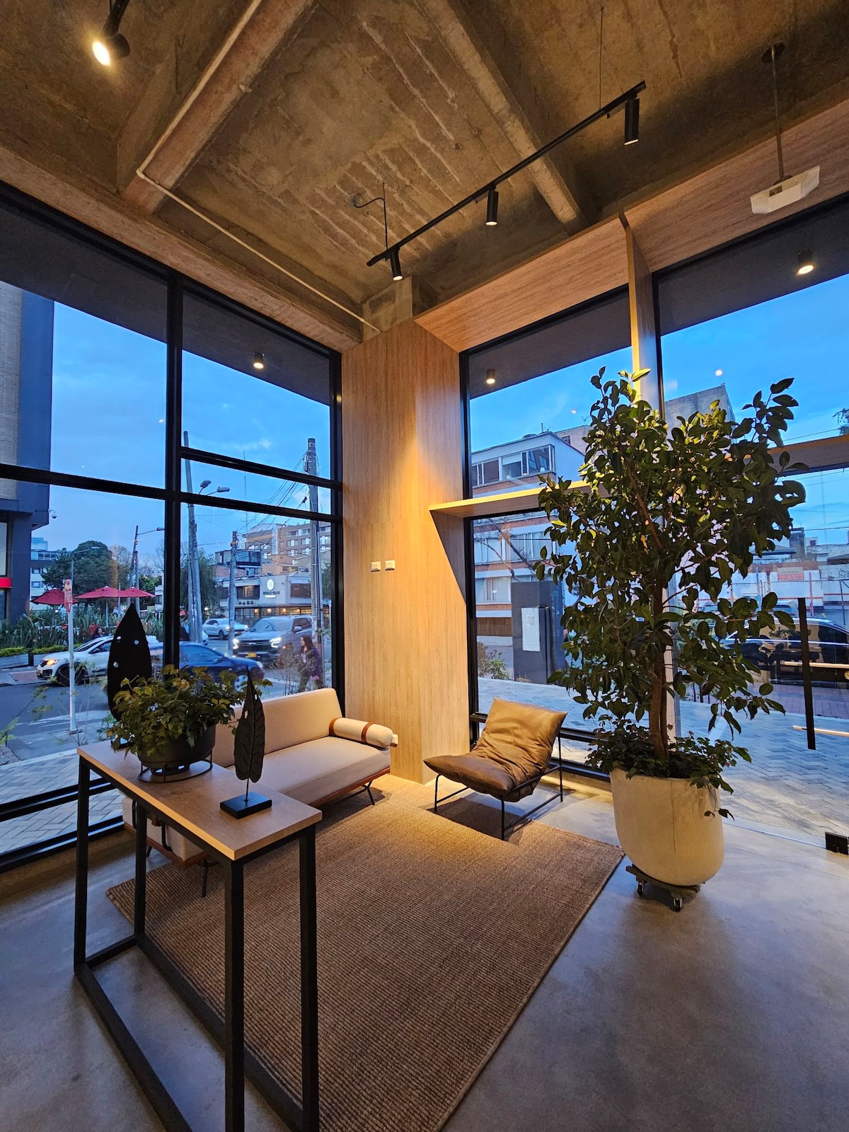 A modern reception area showcases large windows allowing natural light to filter in. Indoor plants add a touch of greenery, while a minimalist table and seating create a welcoming atmosphere. The concrete ceiling contrasts with the wooden accents, providing a stylish and contemporary aesthetic.