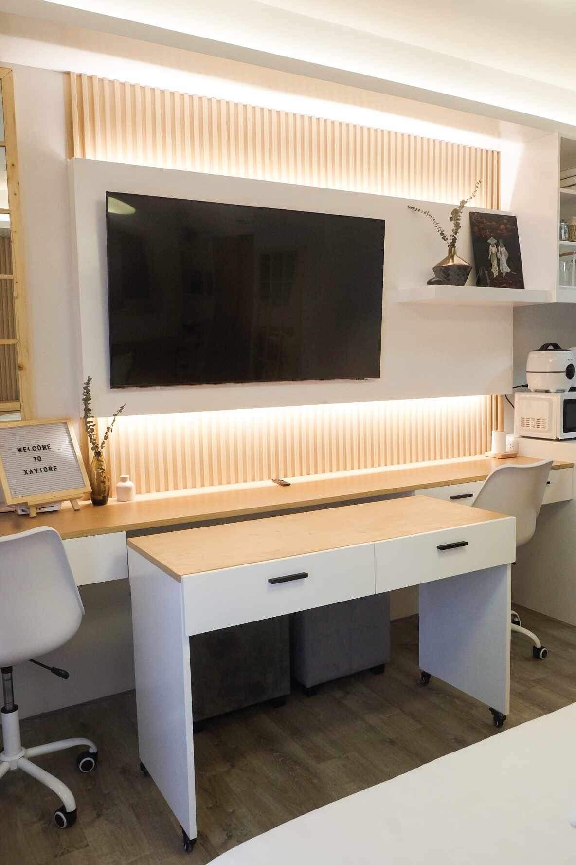 A modern workspace features a light wood desk with two drawers, positioned beneath a large wall-mounted TV. Soft ambient lighting highlights the textured wall above the desk, complemented by minimal decorative items. Two ergonomic white chairs are placed nearby, enhancing the functional design.