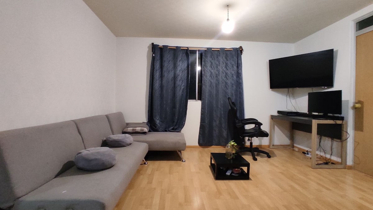 A spacious living area is furnished with a gray couch featuring cushions. A small coffee table occupies the center of the room, accompanied by a chair and desk with a computer setup. Dark curtains frame the window, and a television is mounted on the wall.