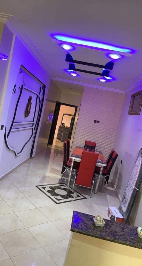 A modern dining area is showcased, featuring a rectangular table surrounded by six sleek, red chairs. The glossy tiled floor reflects light from stylish ceiling fixtures, and a decorative rug lies beneath the table. The walls are accented with subtle patterns and soft lighting.