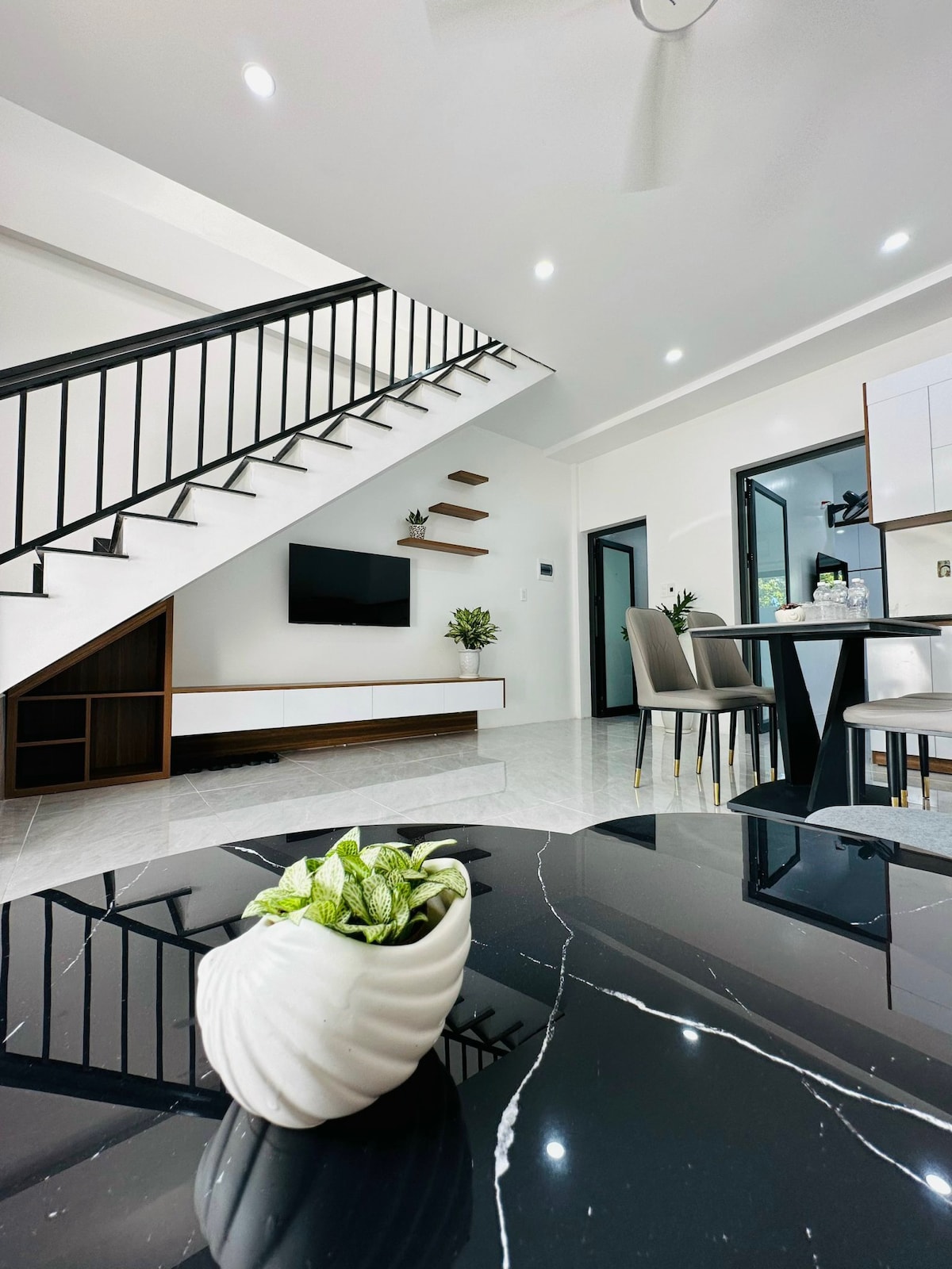A modern living space is displayed, featuring a sleek black marble table with a decorative plant in a white vase. A staircase with a minimalist railing is visible, along with a flat-screen TV mounted on the wall. The open floor plan enhances the spaciousness.