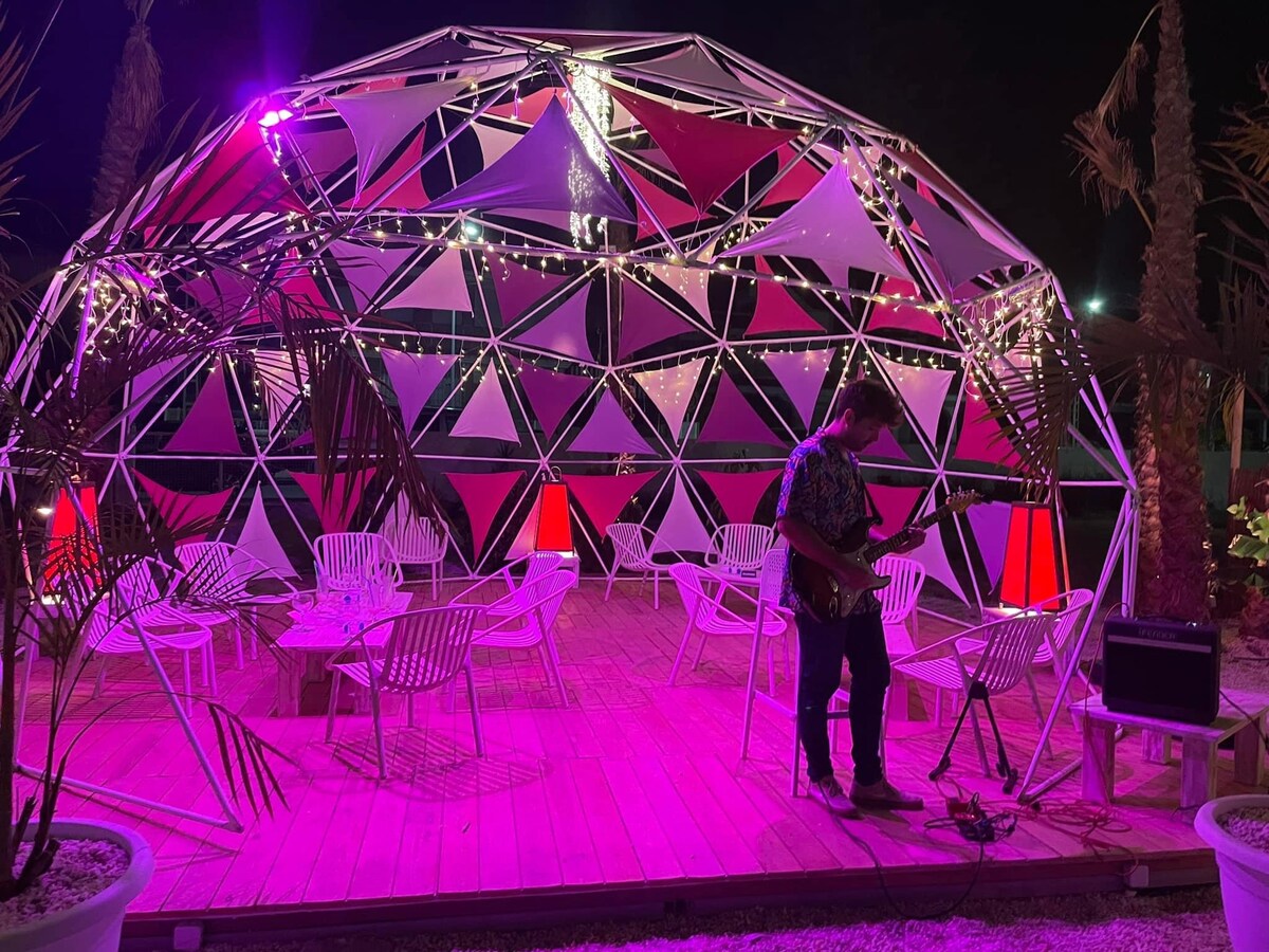 A geometric structure stands illuminated at night, surrounded by vibrant pink and purple lights. Seating is arranged around a wooden deck, creating a relaxed atmosphere. A musician is seen playing guitar, with decorative fabric triangles adding to the lively ambiance.