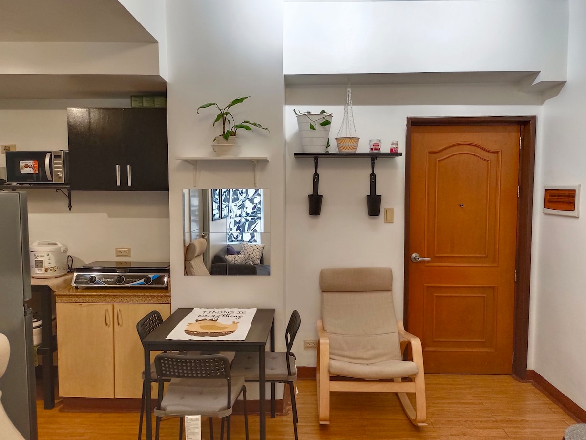 A modern kitchenette is visible with essential appliances, including a microwave and stove. A small dining table with four chairs is positioned nearby. An inviting seating area features a light-colored lounge chair. Natural light enters through the entrance door, enhancing the space's brightness.