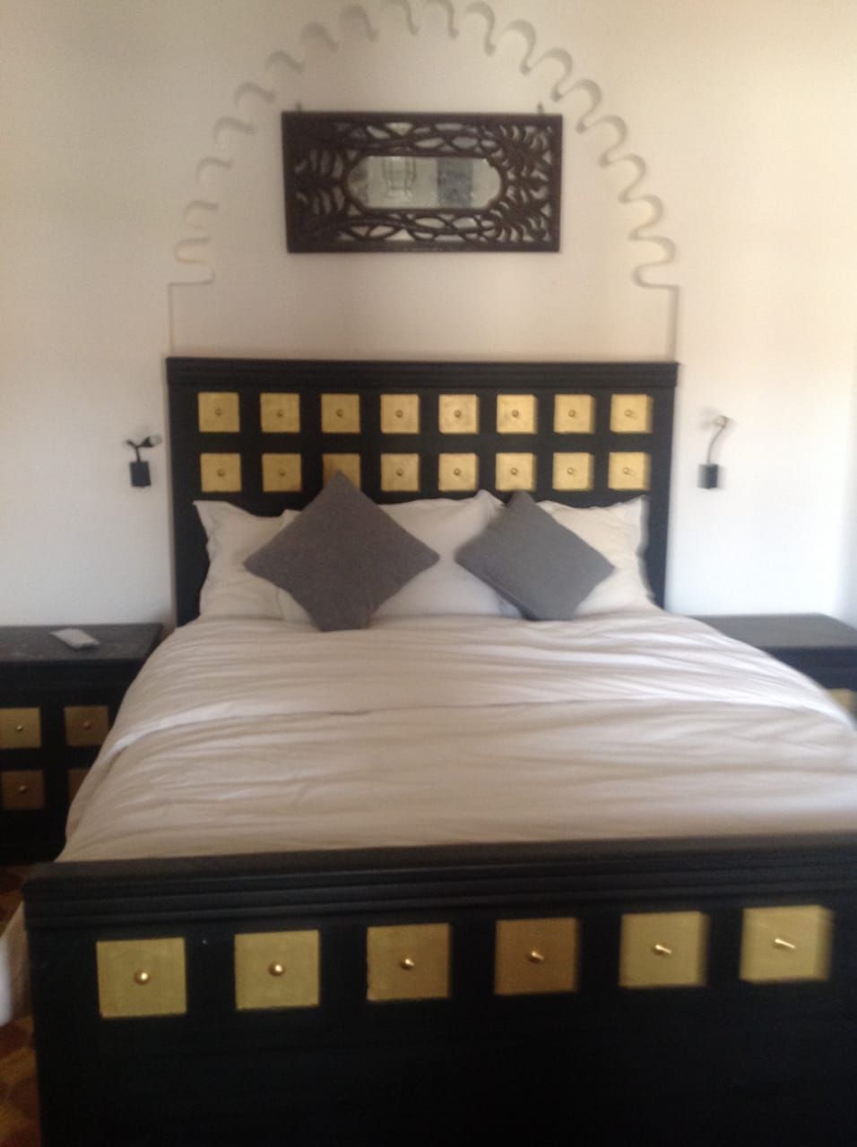 A well-appointed sleeping area features a large bed with decorative gold accents on the headboard. Two gray pillows rest atop crisp white linens. Symmetrical bedside tables with gold detailing are positioned on either side, with wall-mounted lamps providing soft illumination.
