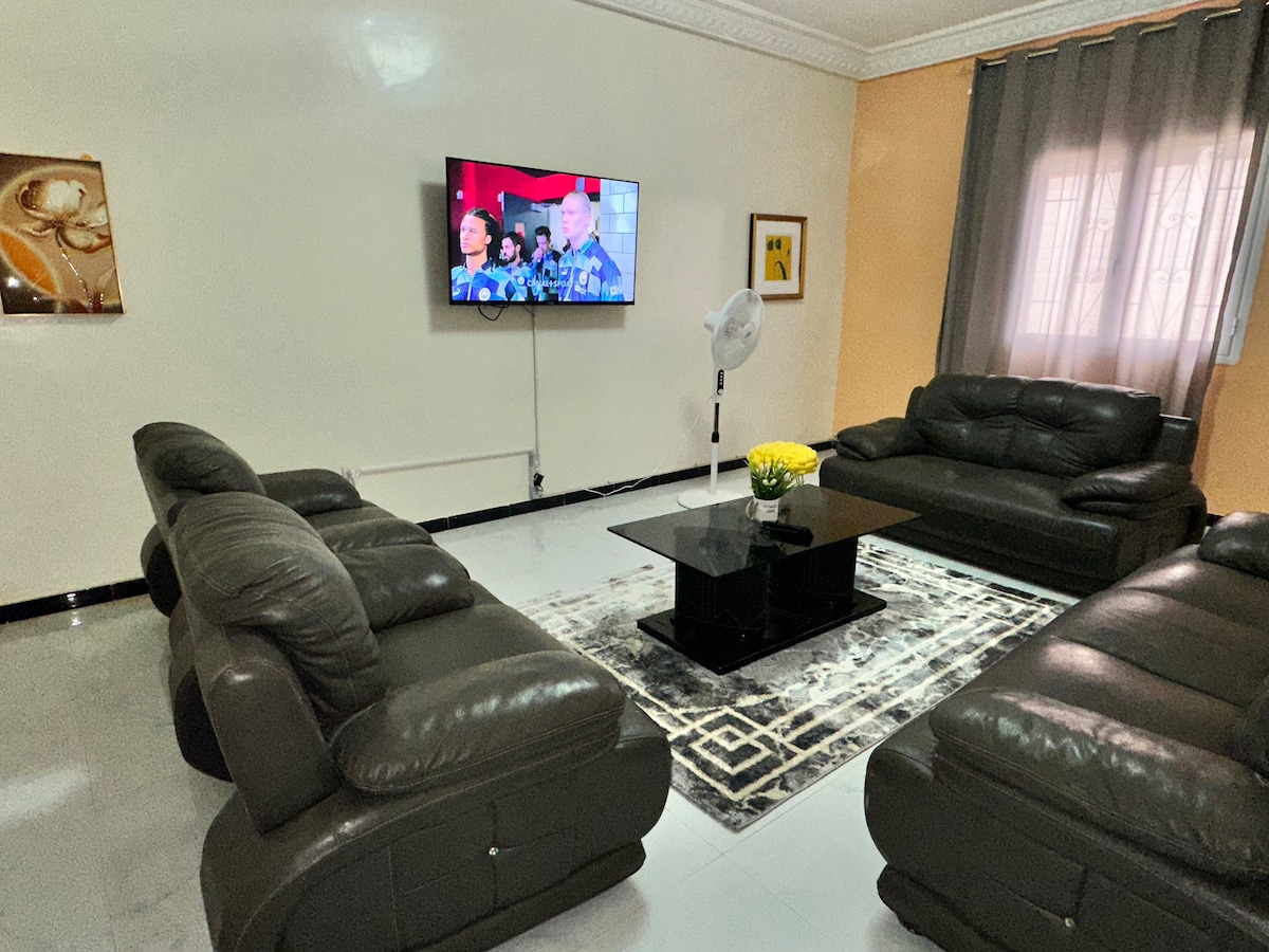 A spacious living area is furnished with black leather sofas arranged around a central glass coffee table. A flat-screen television is mounted on the wall, while a decorative rug adds texture to the polished floor. Natural light filters in through sheer curtains.
