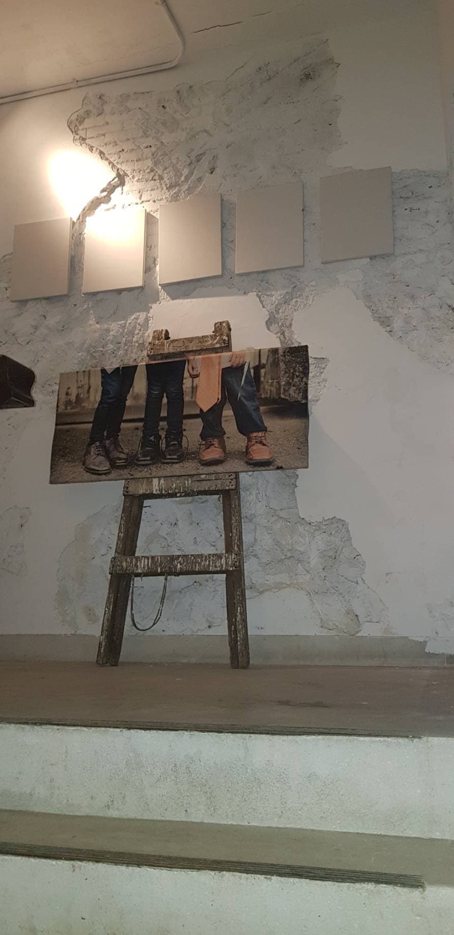 A rustic wooden ladder displays a photograph of individuals standing together, set against a textured wall featuring exposed plaster. Above the photo, several blank canvases are mounted, while soft lighting creates a warm ambiance.