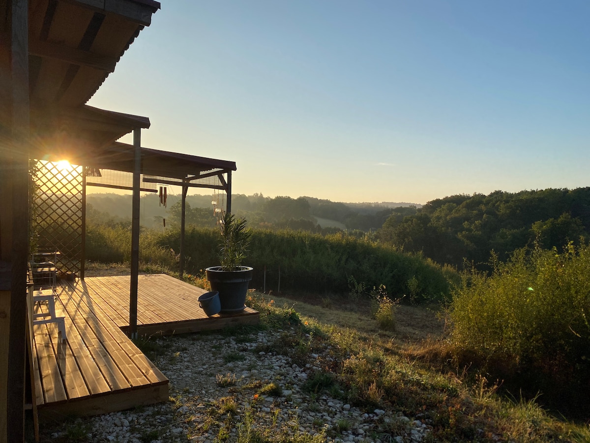 A wooden deck extends from the cabin, with sunlight filtering through a trellis. A large planter sits on the ground, surrounded by greenery and a scenic view of the valley. The landscape features rolling hills and trees under a clear blue sky.