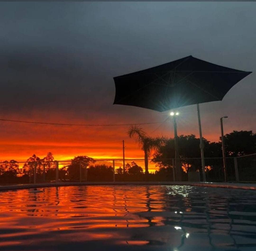 The image captures a pool at sunset, with the vibrant orange and red hues reflecting on the water's surface. A large umbrella provides shade, while soft lighting highlights the area. Silhouettes of trees outline the scene against the dramatic sky.