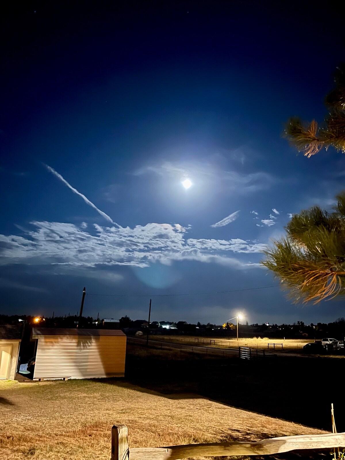 A nighttime scene is captured, featuring a bright moon illuminating the sky above a rural landscape. Wispy clouds are visible, and soft lighting from nearby structures creates a serene atmosphere. The foreground includes silhouettes of trees and an outbuilding against the expansive field.