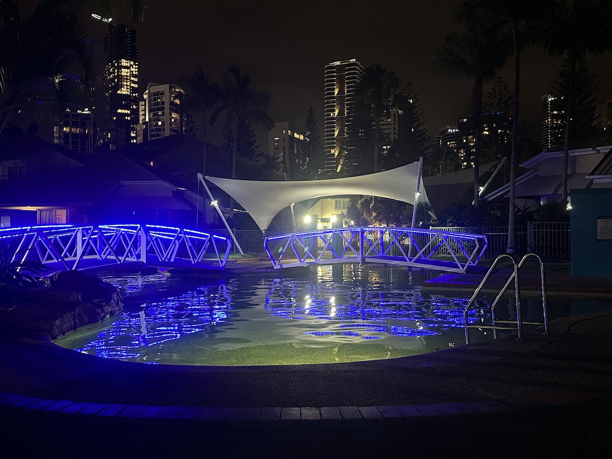 An illuminated lagoon pool is featured, showcasing blue lighting that highlights the water's surface. A fabric canopy provides shade over the pool area. A wooden bridge, adorned with blue lights, spans the pool, leading to lush palm trees and a backdrop of the city skyline.