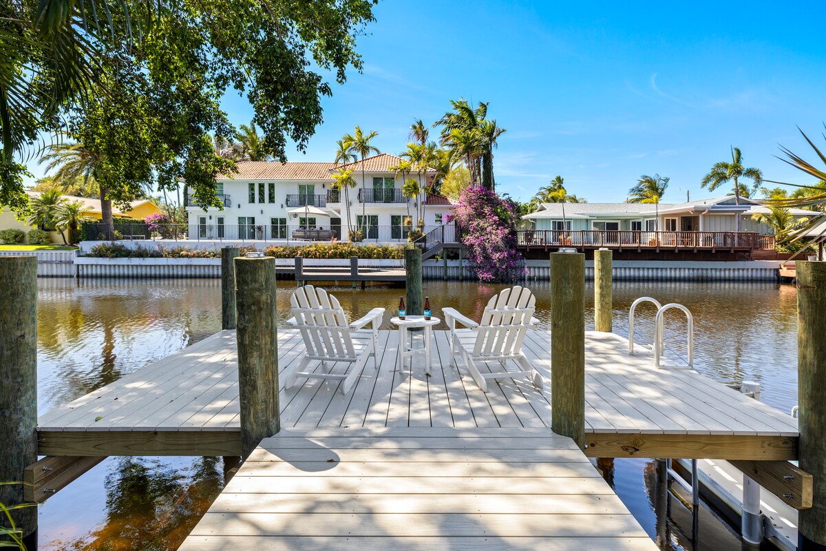 A wooden dock extends over calm waters, flanked by palm trees and tropical foliage. Two white adirondack chairs face the water, providing a relaxing space. In the background, charming homes can be seen along the opposite shoreline, enhancing the tranquil setting.