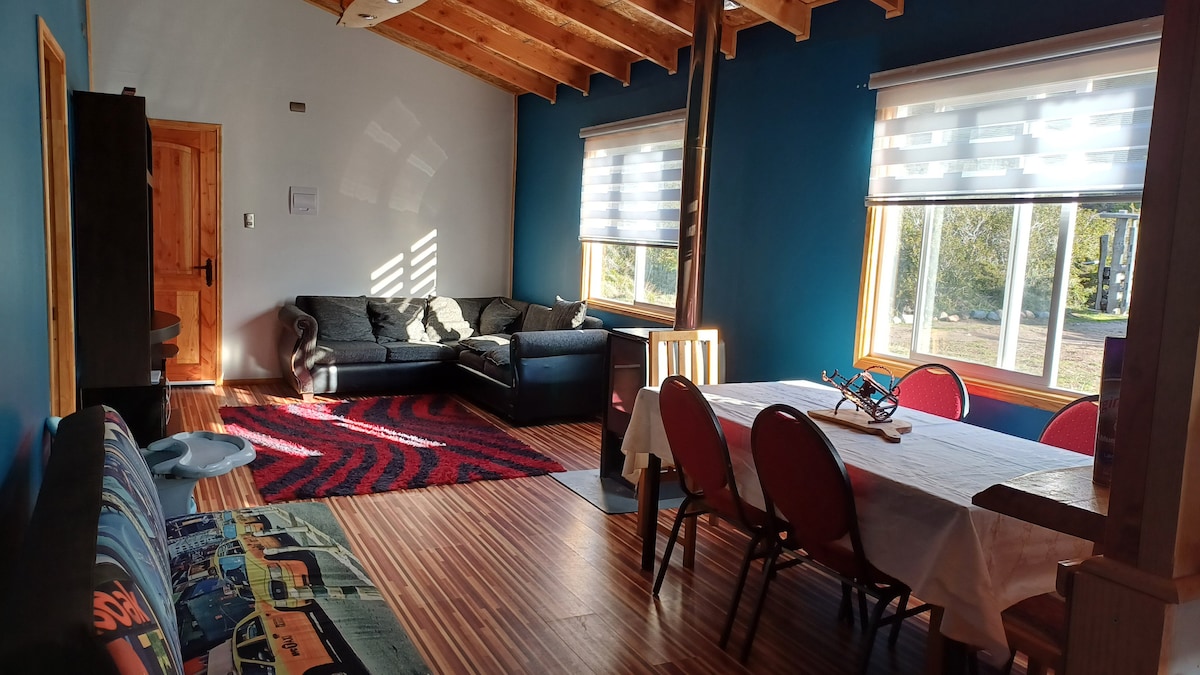 A spacious living area is featured, showcasing a large dining table surrounded by red chairs. Comfortable seating options are arranged, including a dark sofa and a patterned rug. Natural light illuminates the room through large windows with soft roller shades.