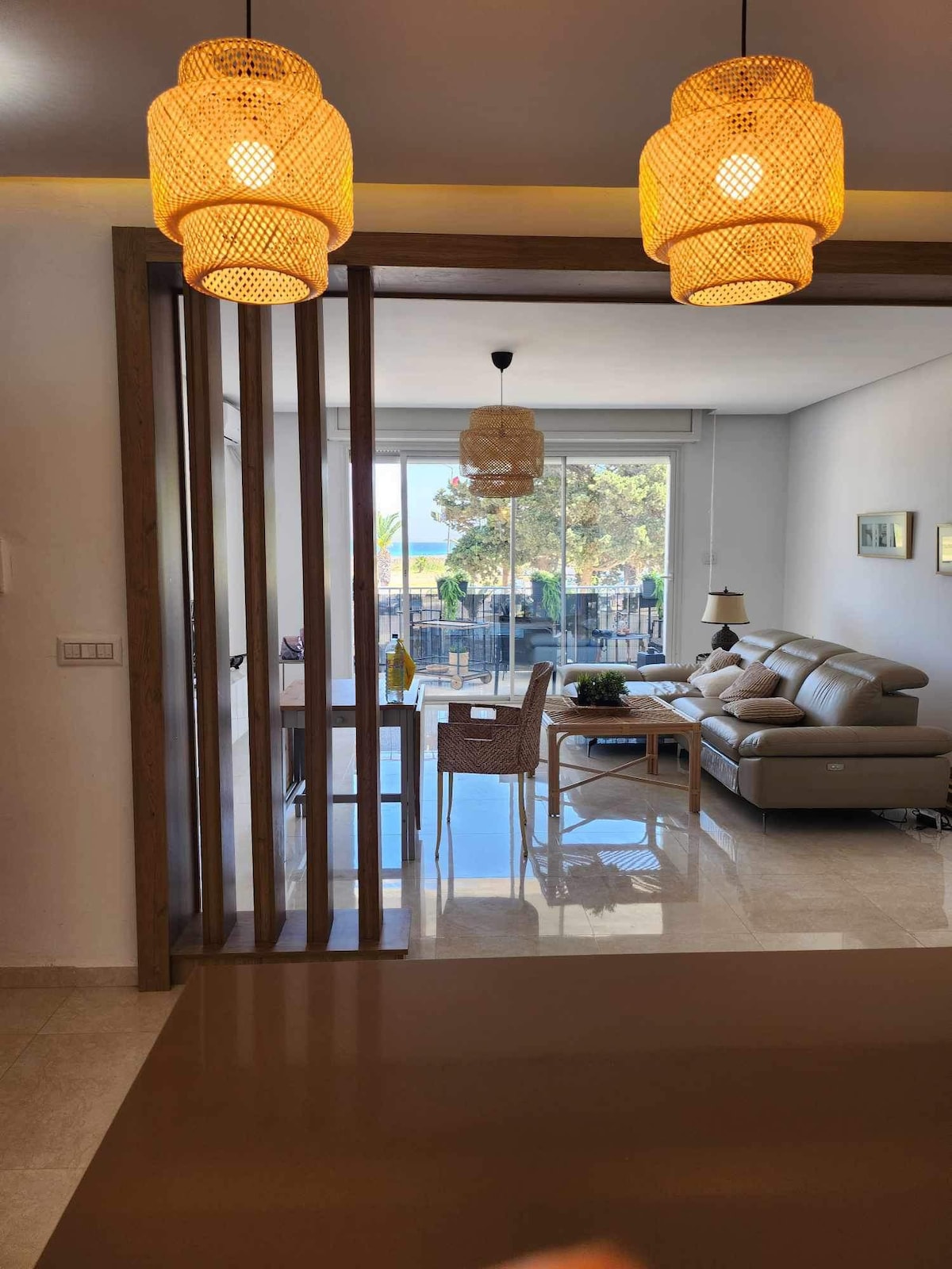 A bright and airy living area is visible through a wooden partition. Comfortable seating is arranged around a coffee table, and large glass doors lead to a sunny outdoor space. Soft lighting fixtures provide warm illumination, enhancing the welcoming atmosphere.