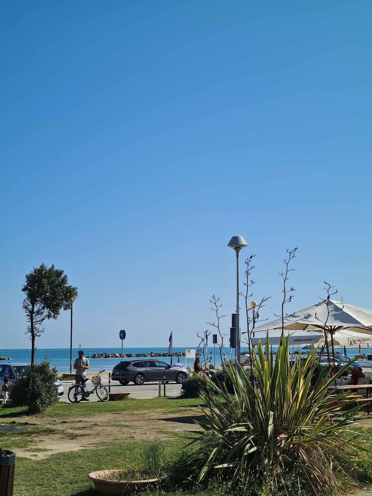 A sunny coastal scene is displayed, featuring a clear blue sky and the ocean in the background. People can be seen enjoying the area, with a cyclist passing by. A well-maintained grassy area is visible, complemented by low shrubs and palm-like plants.