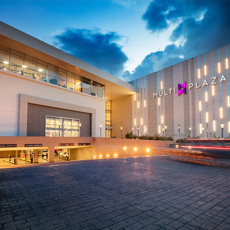 The exterior of the Multiplaza shopping center is displayed, featuring modern architecture with a large illuminated sign. The entrance presents a spacious layout with clean lines, while the parking area below is partially visible. Soft evening light enhances the inviting atmosphere of the setting.