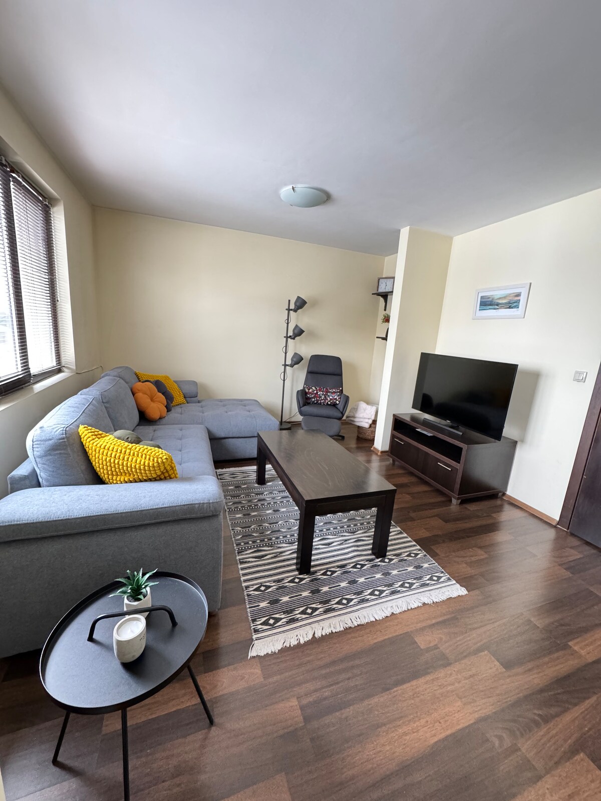 A comfortable living area is furnished with a sectional sofa adorned with yellow accent pillows, a sleek coffee table, and a modern floor lamp. A wall-mounted TV is positioned across from the seating, complemented by a decorative rug on polished hardwood flooring.