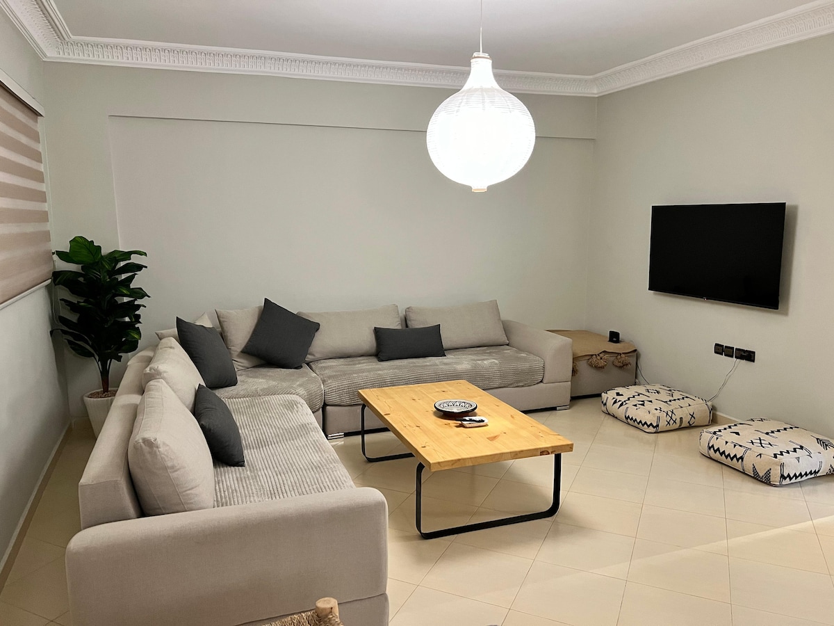 A comfortable living area is presented, featuring a spacious gray sectional sofa adorned with decorative cushions. A wooden coffee table sits at the center, complemented by a flat-screen television mounted on the wall. A plant adds a touch of greenery, enhancing the serene atmosphere.
