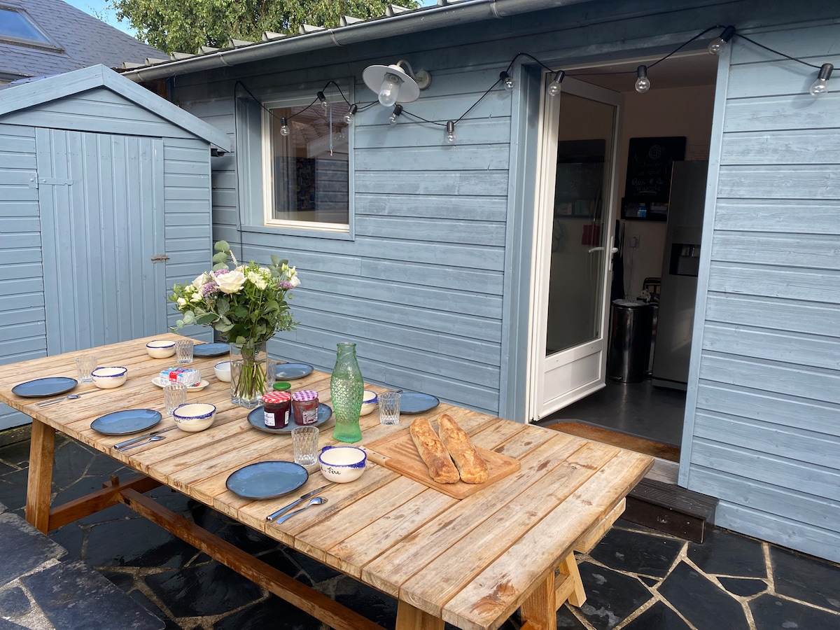 An outdoor dining area is shown with a large wooden table set for a meal. Plates, glasses, and a centerpiece of flowers are arranged on the table. A small shed and a glass door to the interior are visible, complemented by string lights above.
