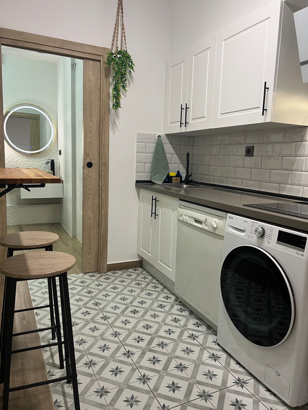 A modern kitchen is presented, featuring stylish white cabinetry and a patterned floor. Essential appliances, including a washing machine and dishwasher, are visible. A small dining area with three stools sits adjacent to the kitchen, while a door leads to a nearby bathroom, enhancing convenience.