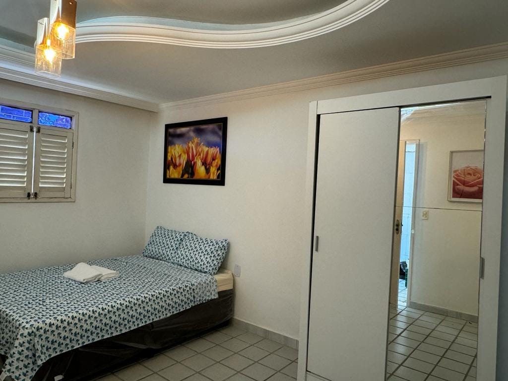 A bright bedroom features a spacious bed dressed in blue and white patterned bedding. A framed floral artwork decorates the wall above the bed. A mirrored wardrobe reflects the room's light, and a portion of the adjoining bathroom is visible through an open door.