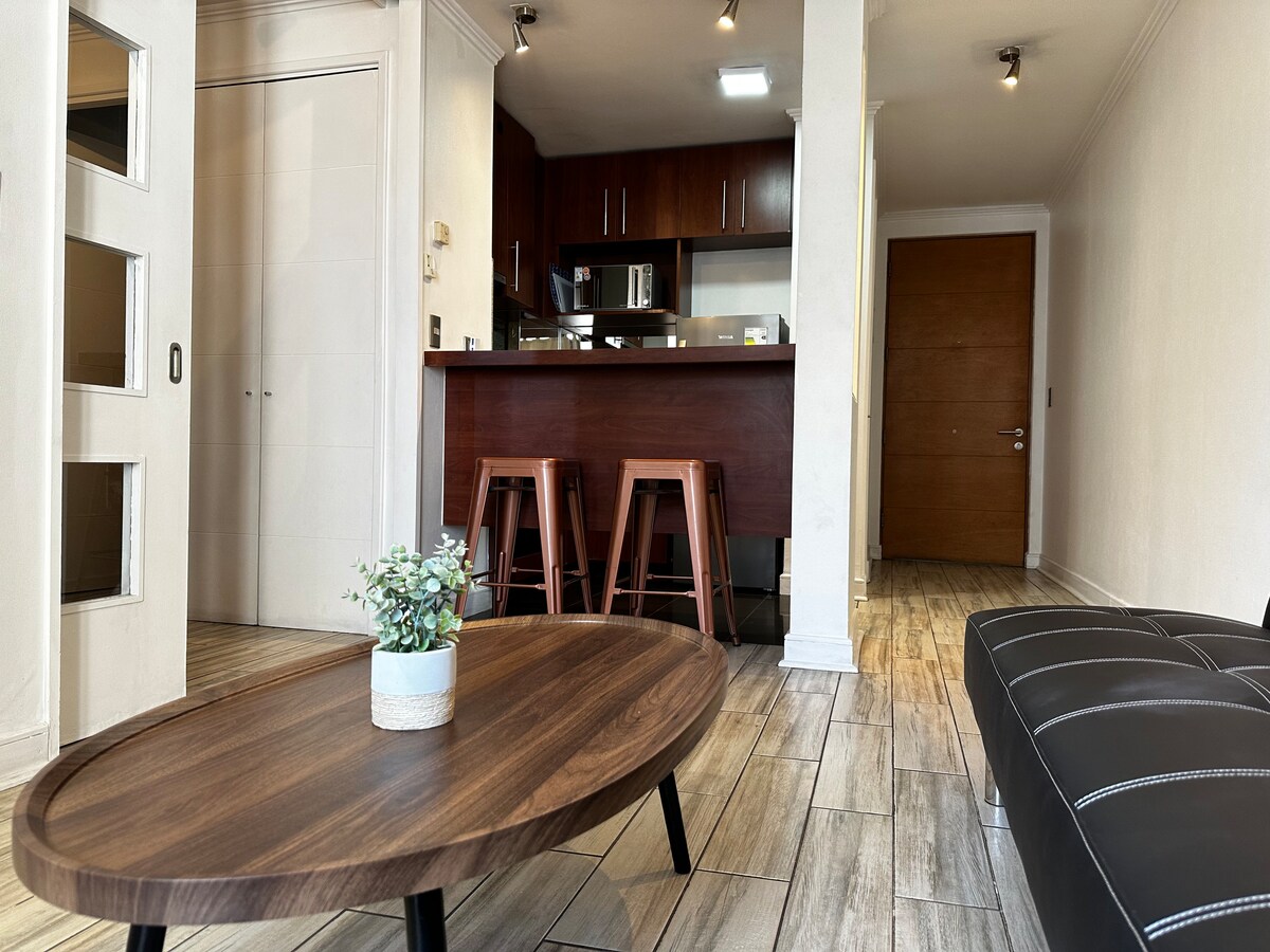 A modern living space features a round wooden coffee table at the center, surrounded by a black sofa. A kitchen bar with three high stools is visible in the background, along with cabinets and appliances. Natural light brightens the space through the open layout.
