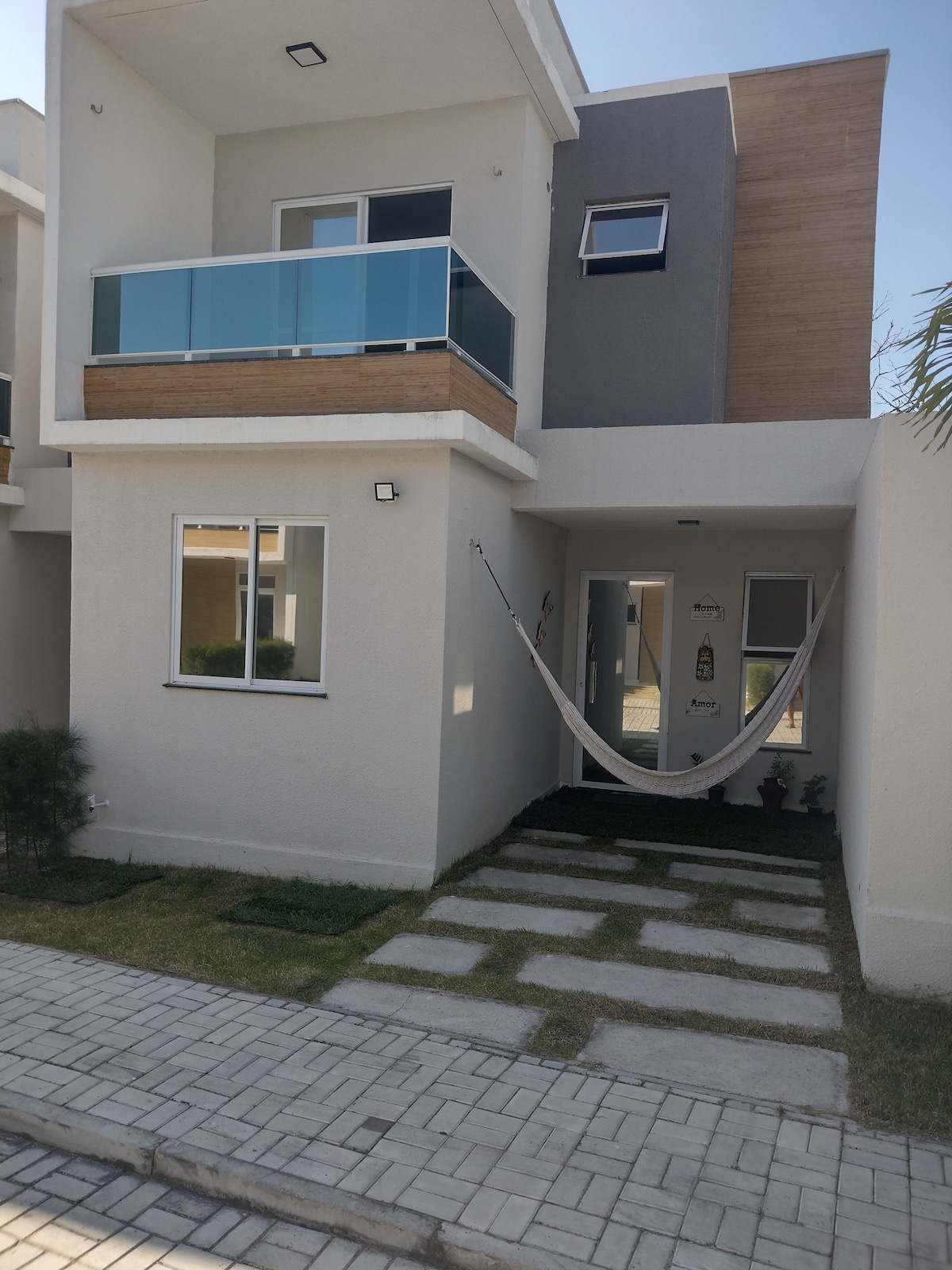 A modern duplex exterior is featured, showcasing a light-colored facade with wooden accents. The entrance is accessed via a pathway lined with stones and grass. A spacious balcony is visible above, with a hammock hanging near the front door, inviting relaxation.