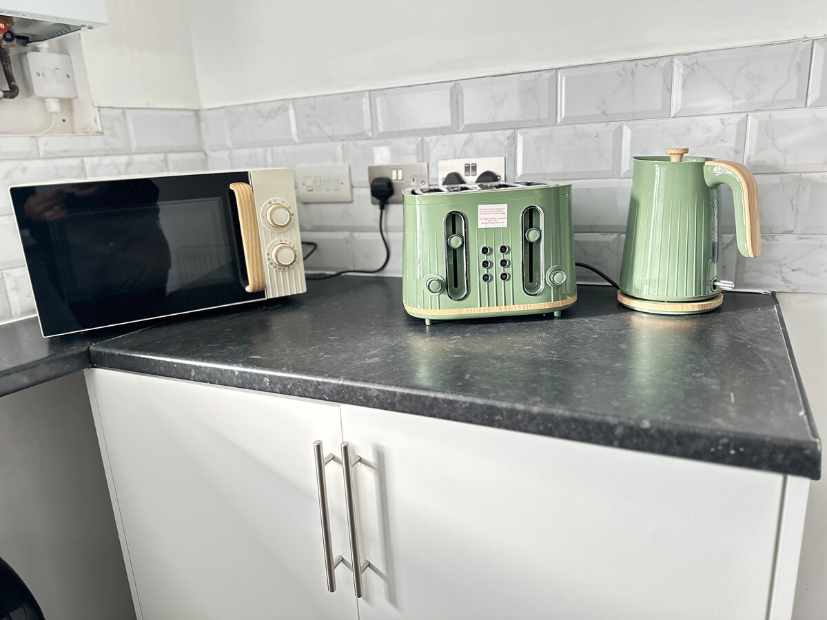 A modern kitchen countertop is shown with a black microwave, a green kettle, and a matching green toaster. The glossy backsplash features light grey tiles, complementing the clean, white cabinetry beneath the countertop.