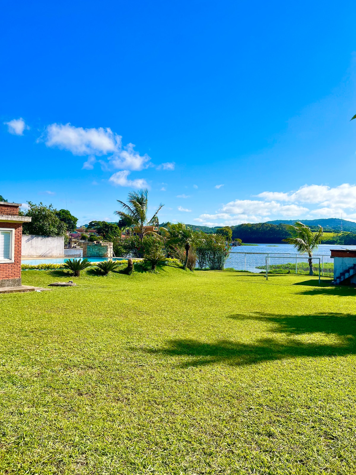 A spacious grassy area is complemented by scattered palm trees and a clear view of the water. The blue sky with a few clouds enhances the serene atmosphere, while nearby structures are visible, providing context to the outdoor space.