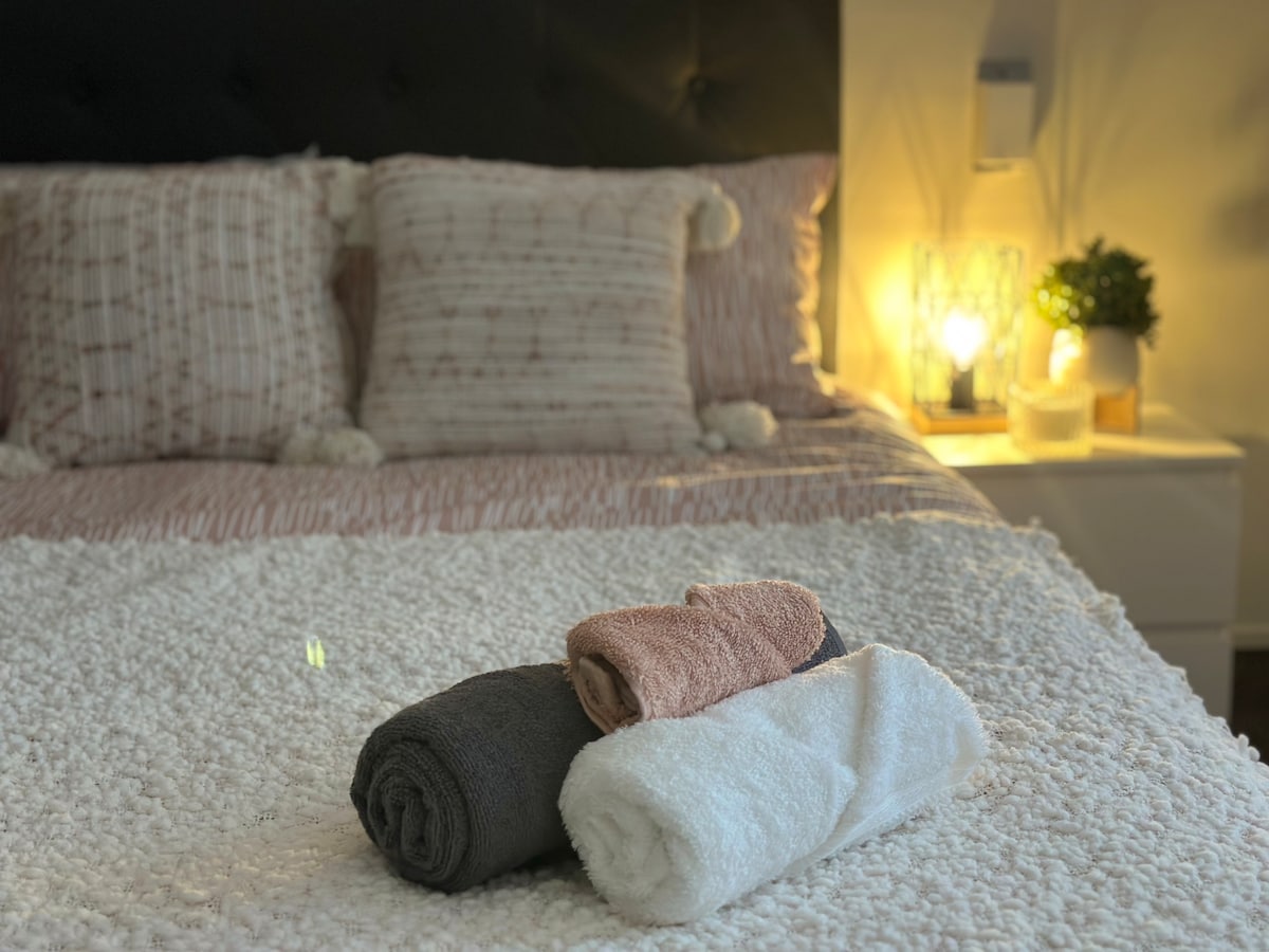 A neatly arranged bed features soft, textured linens and decorative pillows. Three towels, in shades of gray, peach, and white, are placed on a plush, light-colored blanket. A softly glowing lamp and a small plant add a gentle ambiance to the room.