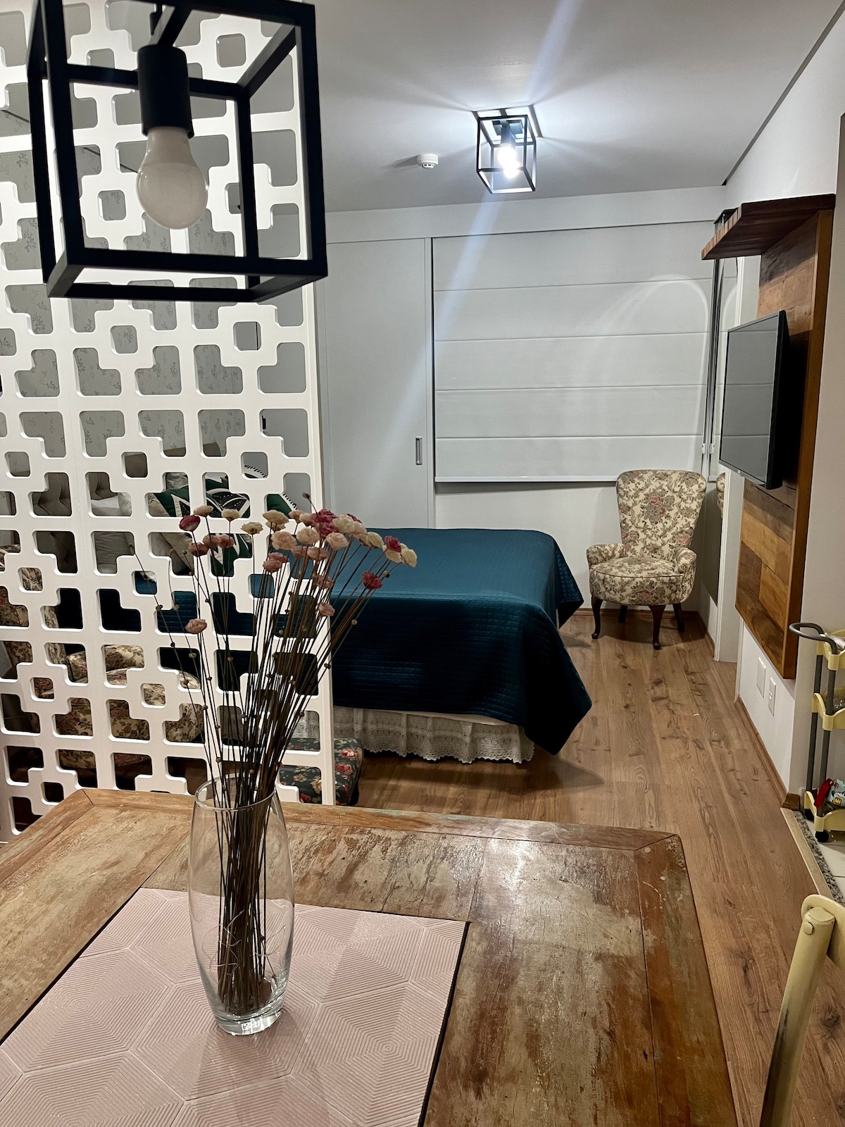 A spacious room is seen, featuring a queen-sized bed draped in a dark blue coverlet. An accent chair is situated beside the bed. A decorative partition separates the sleeping area from a dining space, where a vase with dried flowers is placed on a pink tablecloth.