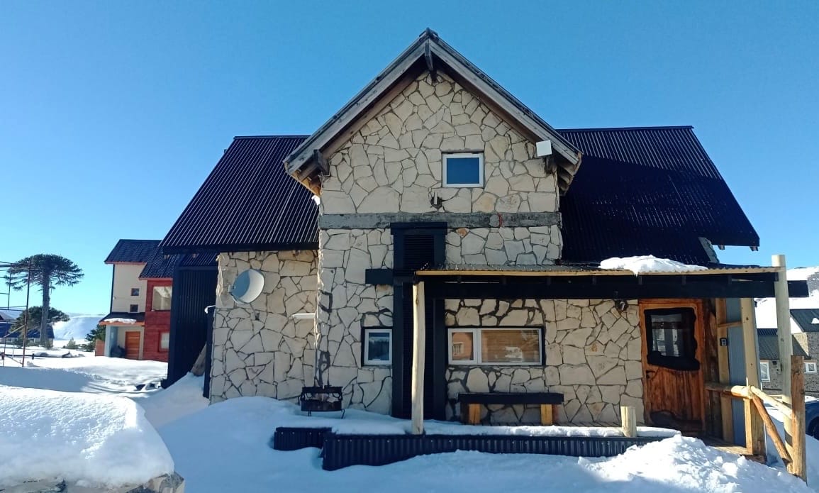 The exterior of the cabin features a sturdy stone facade beneath a sloping roof, complemented by wooden accents. Snow covers the ground, highlighting the rustic charm of the structure. Large windows allow natural light to enter, with a covered porch offering outdoor access.