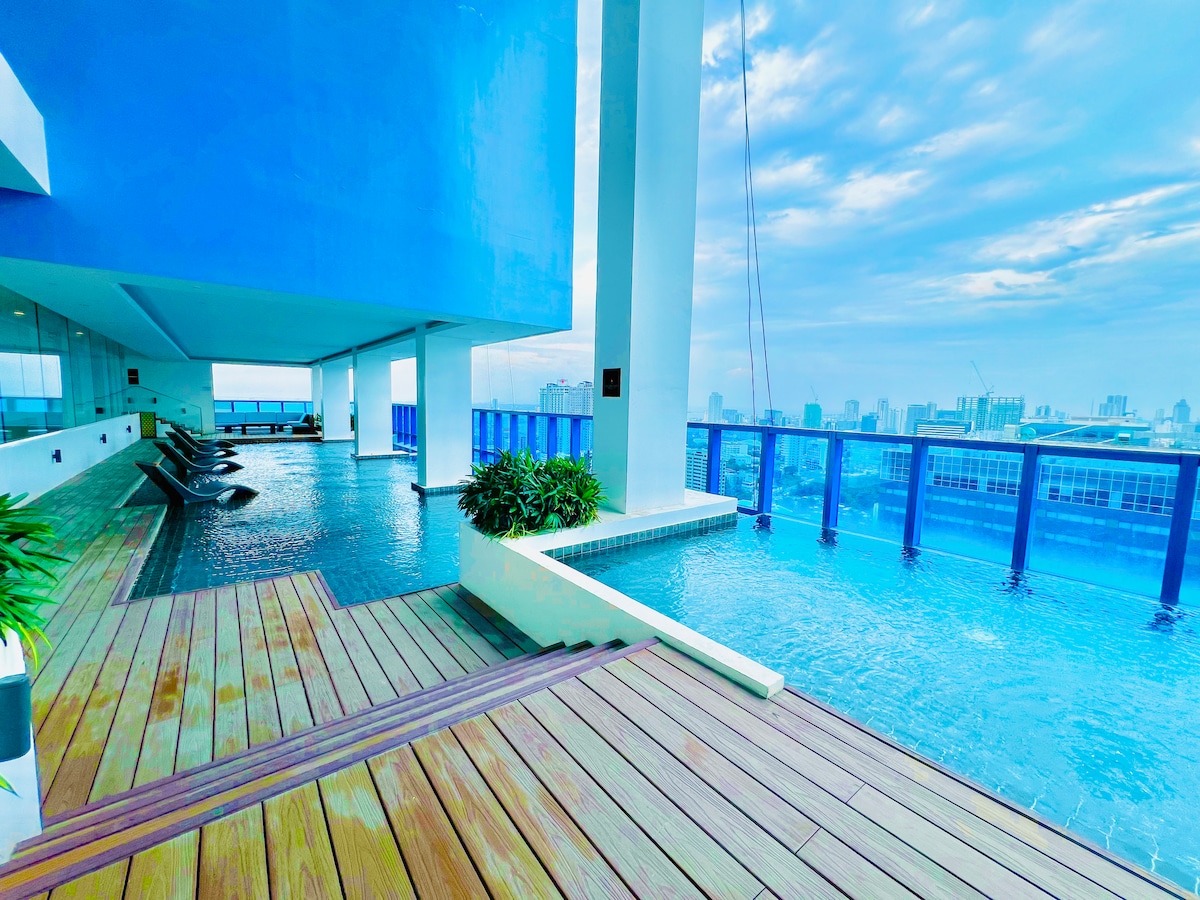 An outdoor pool area features a calm body of water, surrounded by wooden decking and potted plants. Lounge chairs are positioned along the edge, offering a view of the city skyline through transparent glass railings.