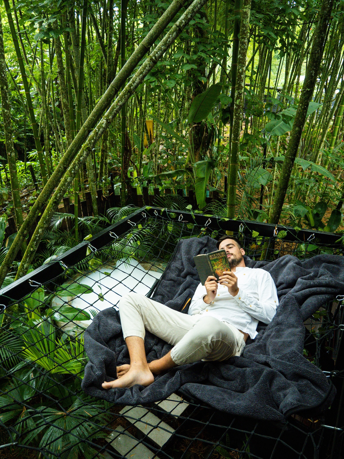 A hammock net is surrounded by lush bamboo and tropical foliage, providing a serene retreat. A blanket is draped over the cozy space, while a person is seen reading a book in a relaxed position, with the natural greenery creating an intimate atmosphere.