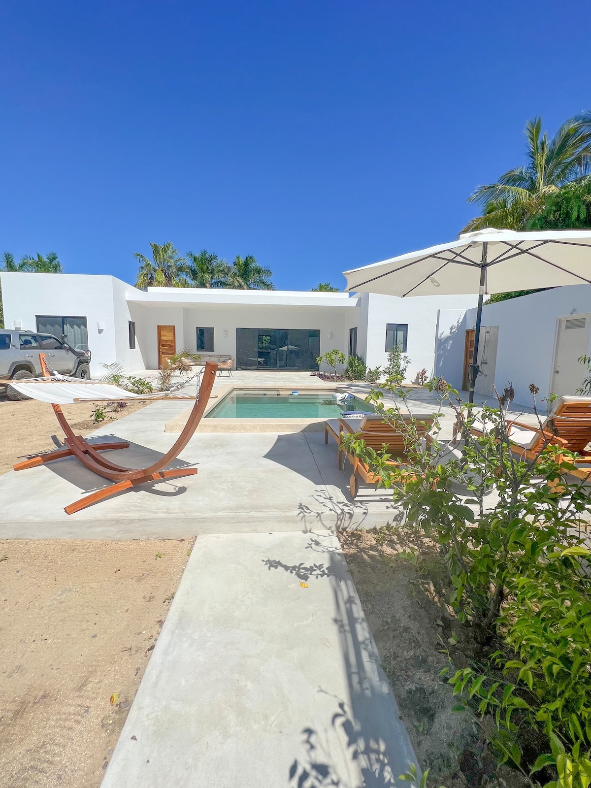 The outdoor area features a shared pool surrounded by a concrete patio, complemented by wooden lounge chairs. A large umbrella provides shade, and several flowering plants enhance the natural setting. The guesthouse stands in the background, set against a clear blue sky.