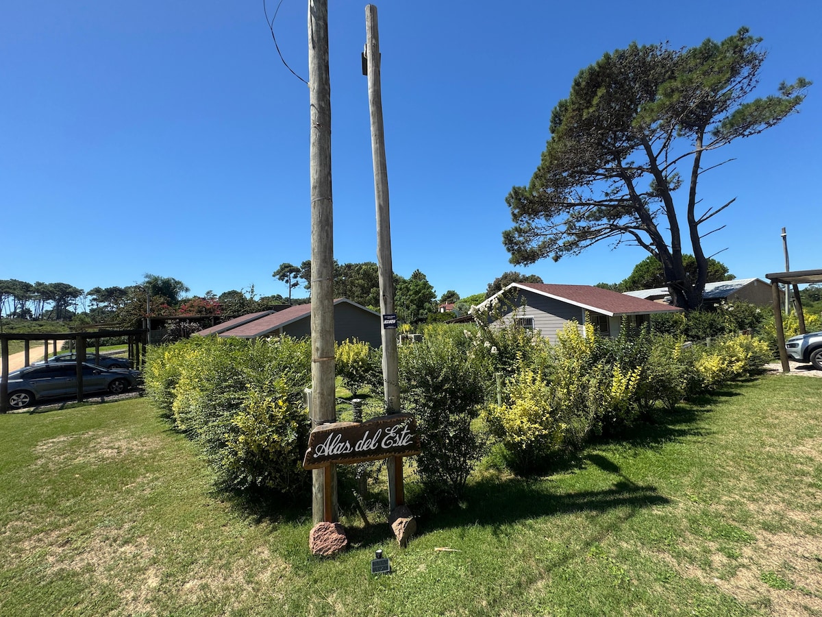 The entrance to Alas del Este is marked by a wooden sign set among lush greenery. In the background, several cabins are visible, framing a clear blue sky. Two power poles stand prominently, indicating a welcoming, natural setting for guests.
