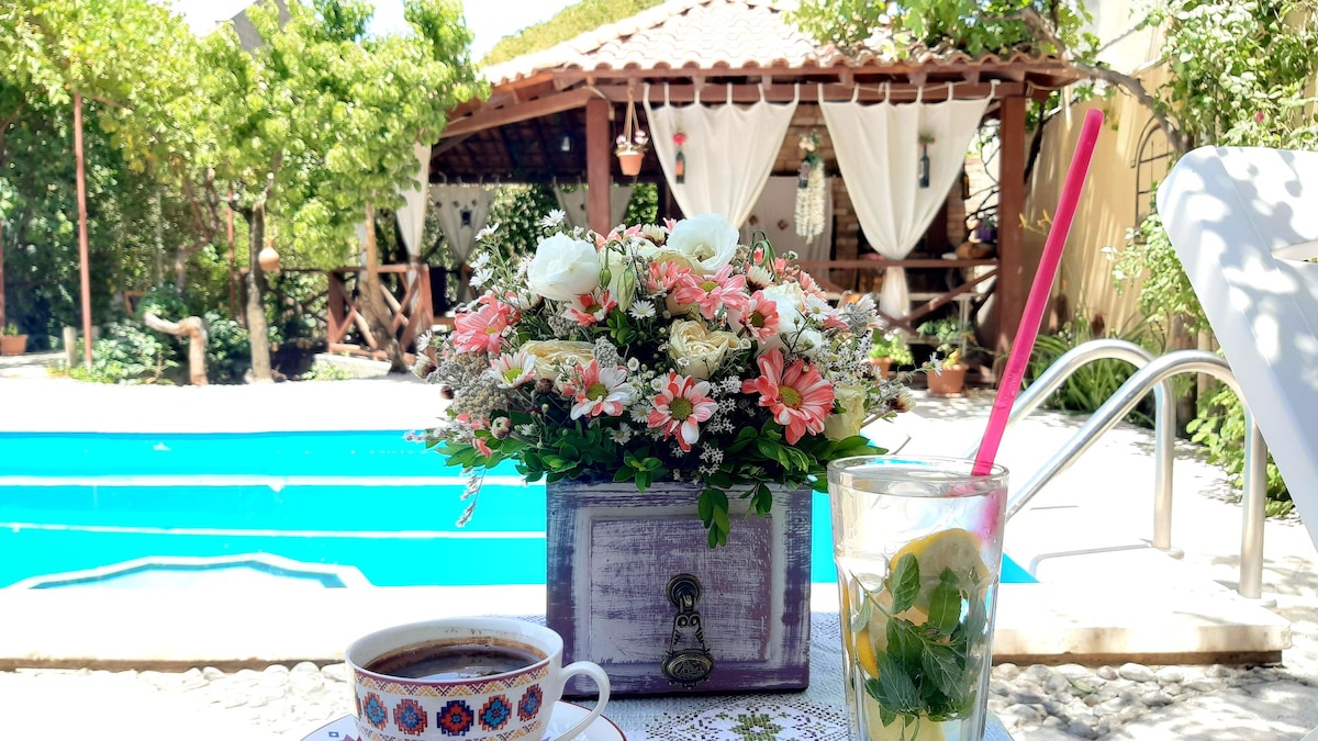A cozy outdoor setting features a beautifully arranged flower box with pink and white blooms, placed next to a cup of coffee and a refreshing drink. In the background, an inviting swimming pool reflects sunlight, and a shaded pavilion is adorned with draped fabric, surrounded by lush greenery.