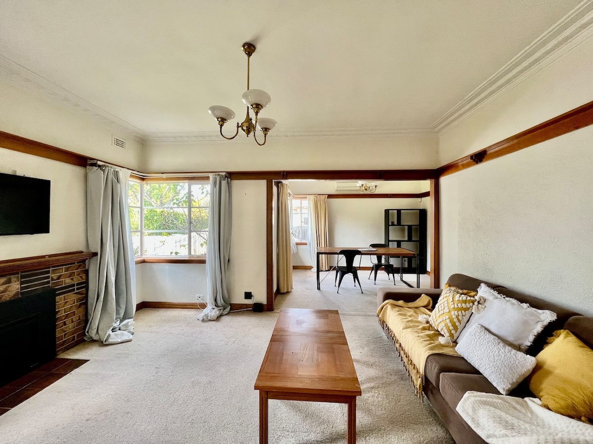 A spacious living area is presented with soft carpeting and natural light filtering through large windows framed by curtains. A comfortable sofa is positioned opposite a decorative fireplace, while a wooden coffee table sits in front. A dining area with a dark table is visible in the background.