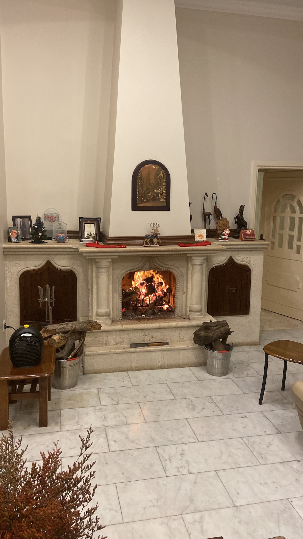 A traditional fireplace is centered within a stone mantel, creating a focal point in the room. Soft flames flicker inside, contrasting with light marble flooring and neutral walls. Decorative items are arranged on the mantel, and a wooden chair is positioned nearby.