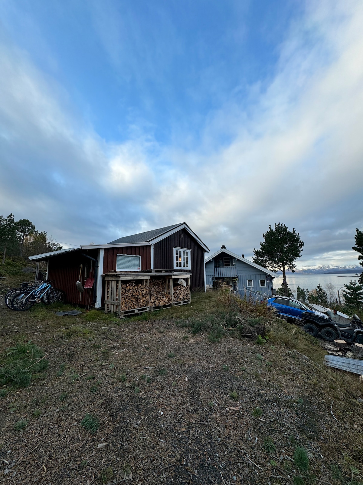 A cozy cabin in natural surroundings features a wood storage area and bicycles against the side. The building's rustic exterior is complemented by a varied color palette, with a spacious view of the landscape visible in the background.