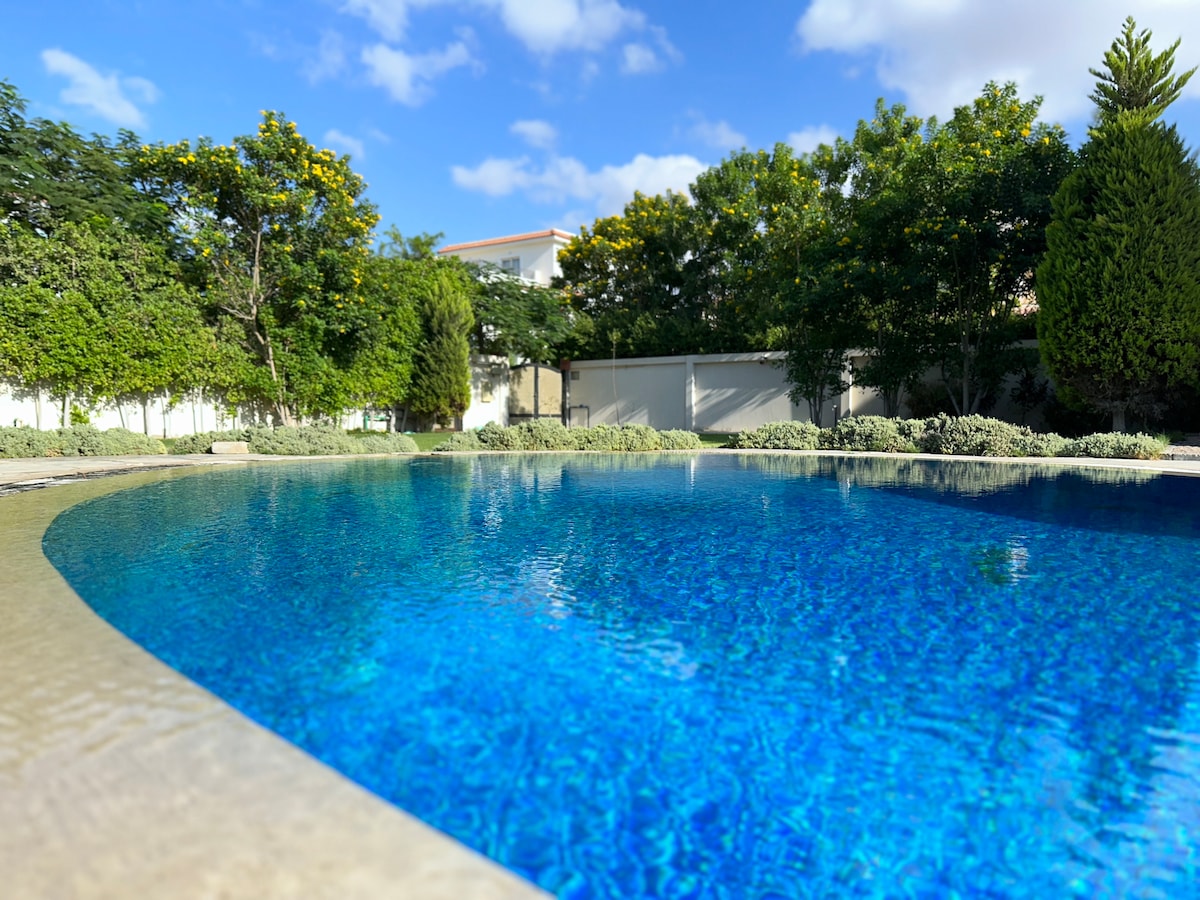 A tranquil swimming pool is surrounded by lush greenery and tall trees. The water shimmers in bright blue under a clear sky, reflecting the natural beauty of the garden area. The pool area is designed for relaxation and enjoyment.