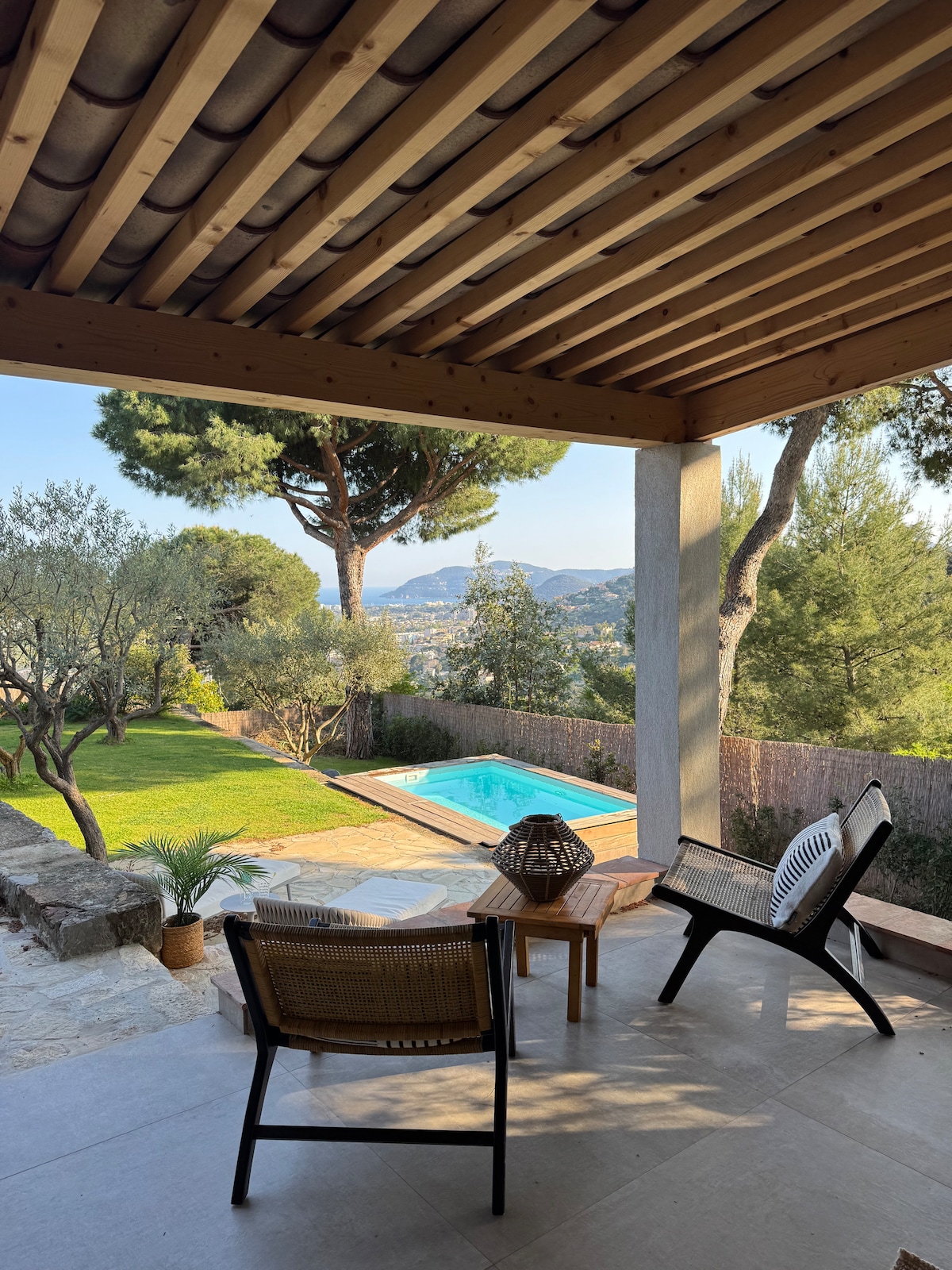 A shaded terrace features two stylish chairs facing a private pool, surrounded by lush greenery. A small table is placed nearby, and distant mountains are visible beyond the well-maintained garden. The scene reflects a peaceful outdoor area ideal for relaxation.