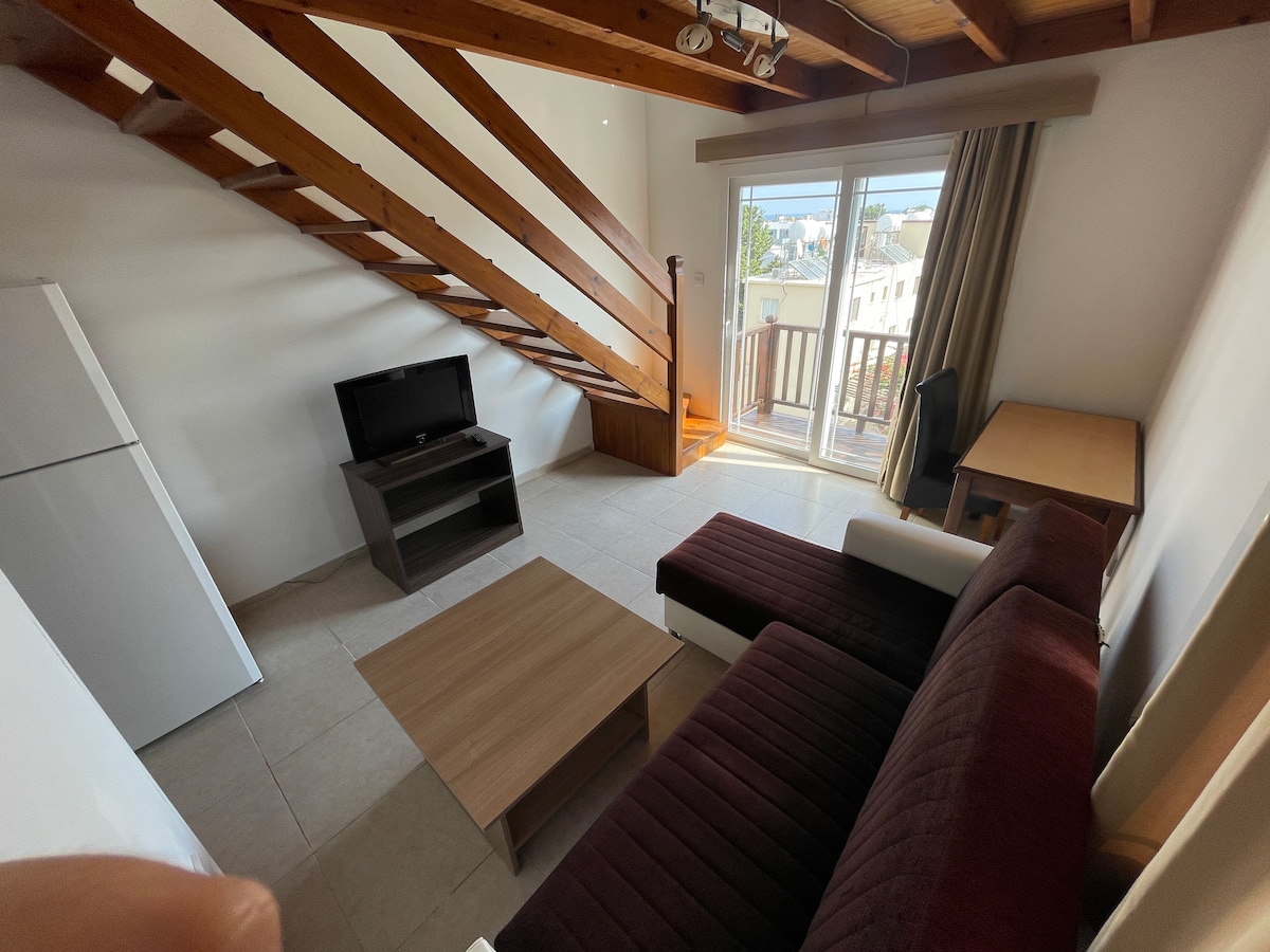 A well-arranged living area is showcased with a brown sofa, a wooden coffee table, and a small desk. Natural light fills the space through sliding glass doors that lead to a balcony. A staircase with wooden steps is visible in the background.