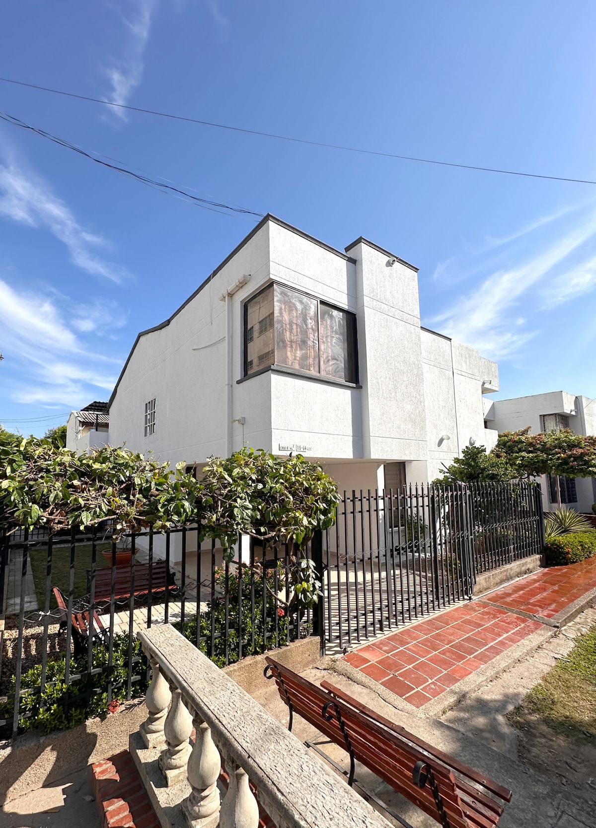 The exterior of a modern two-story house is depicted, featuring white walls and large windows. A well-maintained garden surrounds the property, with paved pathways and decorative benches. A gated entrance provides access to the home, which is bathed in natural sunlight under a clear blue sky.