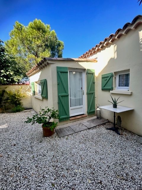 An independent studio is presented, featuring light green shutters and a welcoming entrance door. A small table is set near the entry, surrounded by a gravel area and a variety of greenery, enhancing the outdoor space's natural feel.