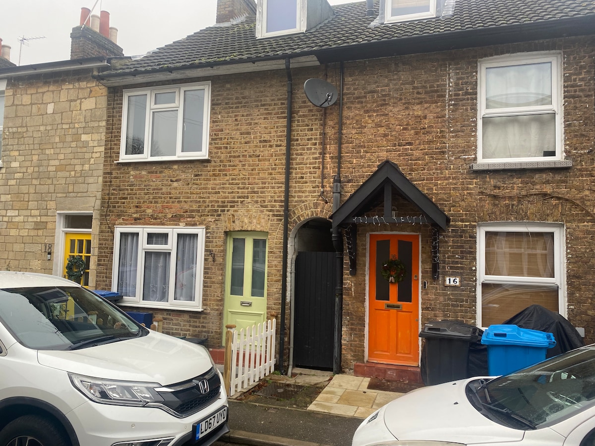 The exterior of a Victorian cottage is displayed, featuring a traditional brick facade. The entrance is highlighted with an orange door, while a yellow door is visible nearby. Vehicles are parked along the street, and a white picket fence is seen beside the green entrance.
