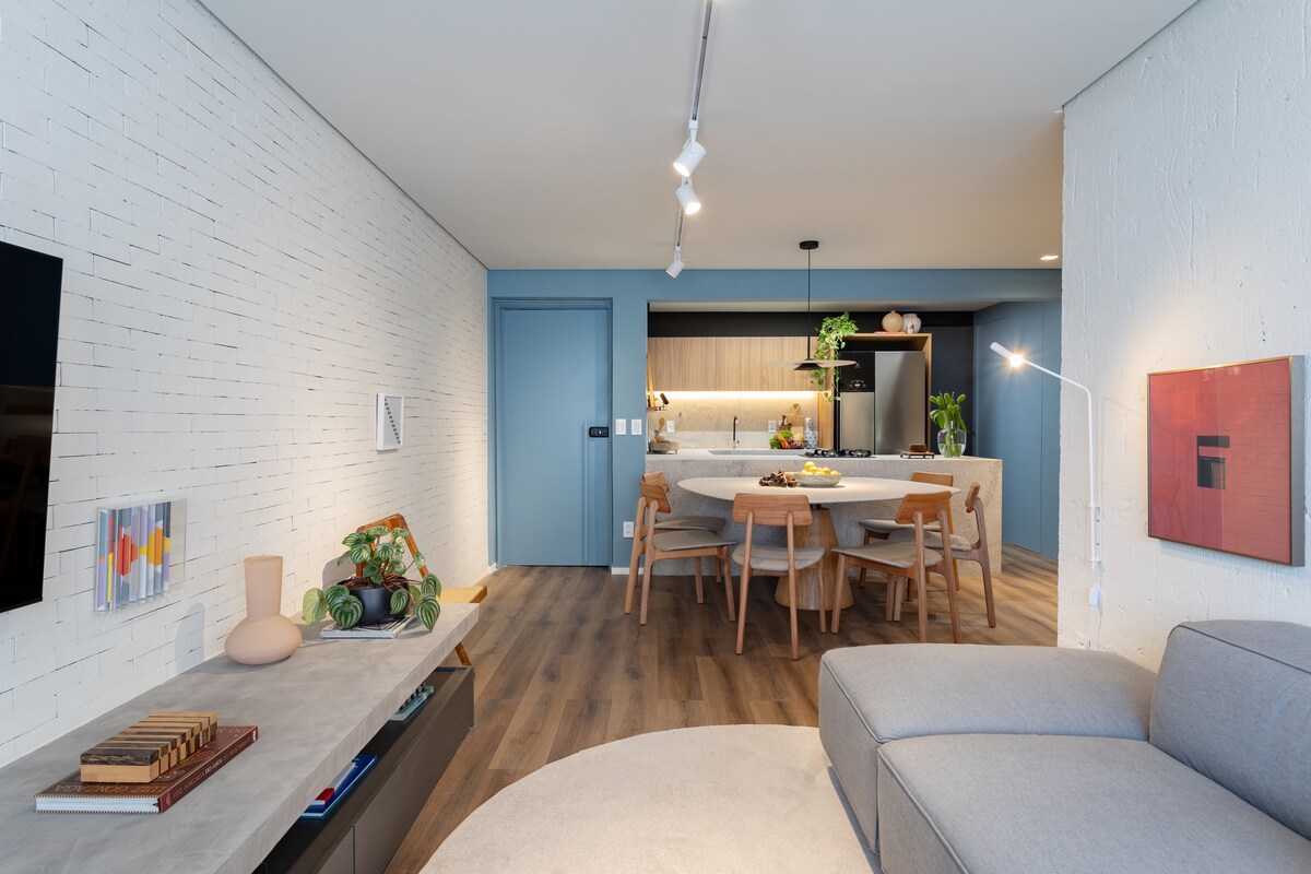 A spacious living area features a light-colored sectional sofa and a round area rug, complemented by wooden flooring. An open kitchen is visible in the background, with modern cabinetry and a dining table surrounded by chairs, creating a welcoming environment for gatherings.